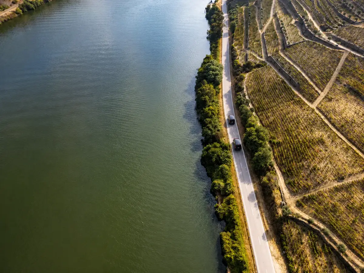 Zwei schwarze Fahrzeuge auf einer Straße entlang des Douro, terrassierte Weinberge am gegenüberliegenden Ufer